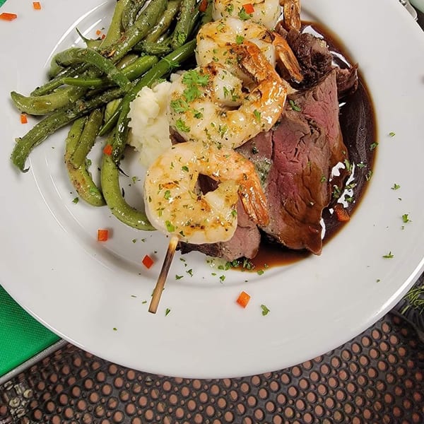 Grilled shrimp skewer, sliced steak with gravy, mashed potatoes, and green beans on a white plate.