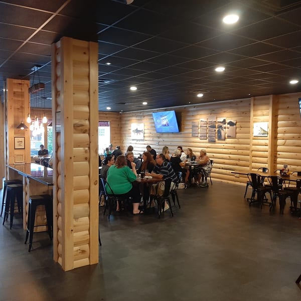 Restaurant interior with wooden walls and columns, people dining at tables, and a bar area.